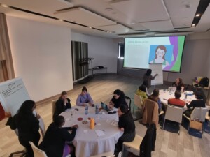 Image shows the participants of the Lupus Europe 2nd Patient Panel on Youth, in a meeting room. Participants are split into two groups, across two tables and they are in the middle of a workshop. A slide on being a young patient with lupus is projected onto the screen. 