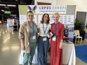 The Lupus Europe booth at EULAR 2023 in Milan. Background of a big Lupus Europe banner that covers the back of the picture. Three women standing in front of the Lupus Europe booth table. The table has leaflets and Lupus100 books. One of the women is holding Lupus100 leaflets. On the left hand side of the picture is the Congress corridor. On background of the right hand side of the picture is a chair